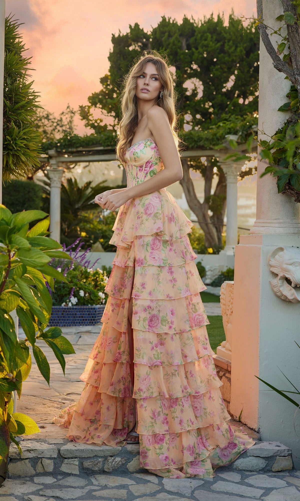 A woman stands outdoors at sunset in the Juliet JT2739S long prom dress, featuring a strapless floral design with a corset bodice and tiered chiffon A-line skirt, surrounded by greenery and white stone structures.